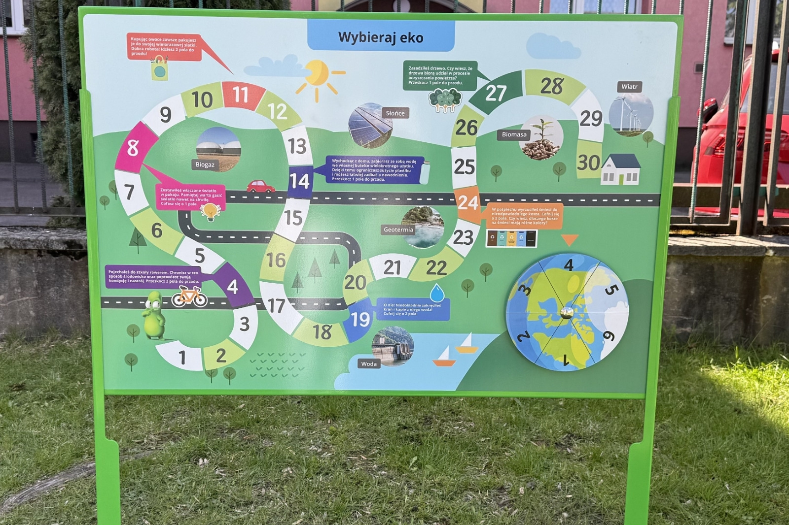 Tablica z grą EKO na dwóch zielonych słupkach - cała plansza z krętą ścieżką pól i obrotową tarczą w prawym dolnym rogu